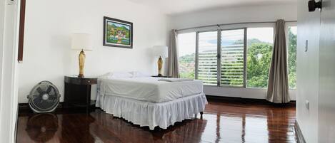 Ein Hotelzimmer mit Bett, zwei Nachttischlampen, einem Standventilator, einem großen Fenster mit Jalousien und einem Gemälde an der Wand.