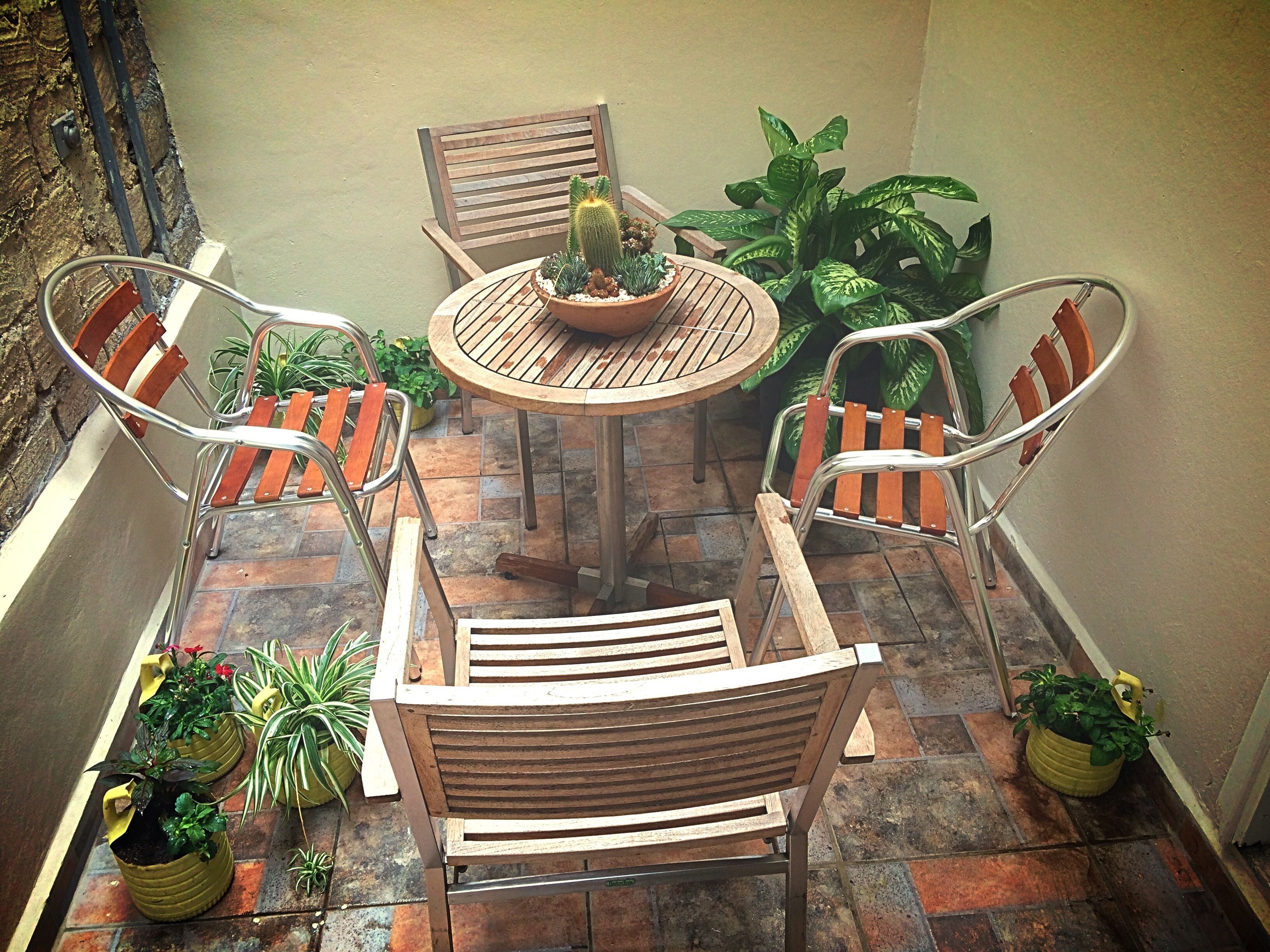 Un petit espace patio avec une table ronde et quatre chaises en bois, entouré de plantes en pot et d’une autre chaise en bois.