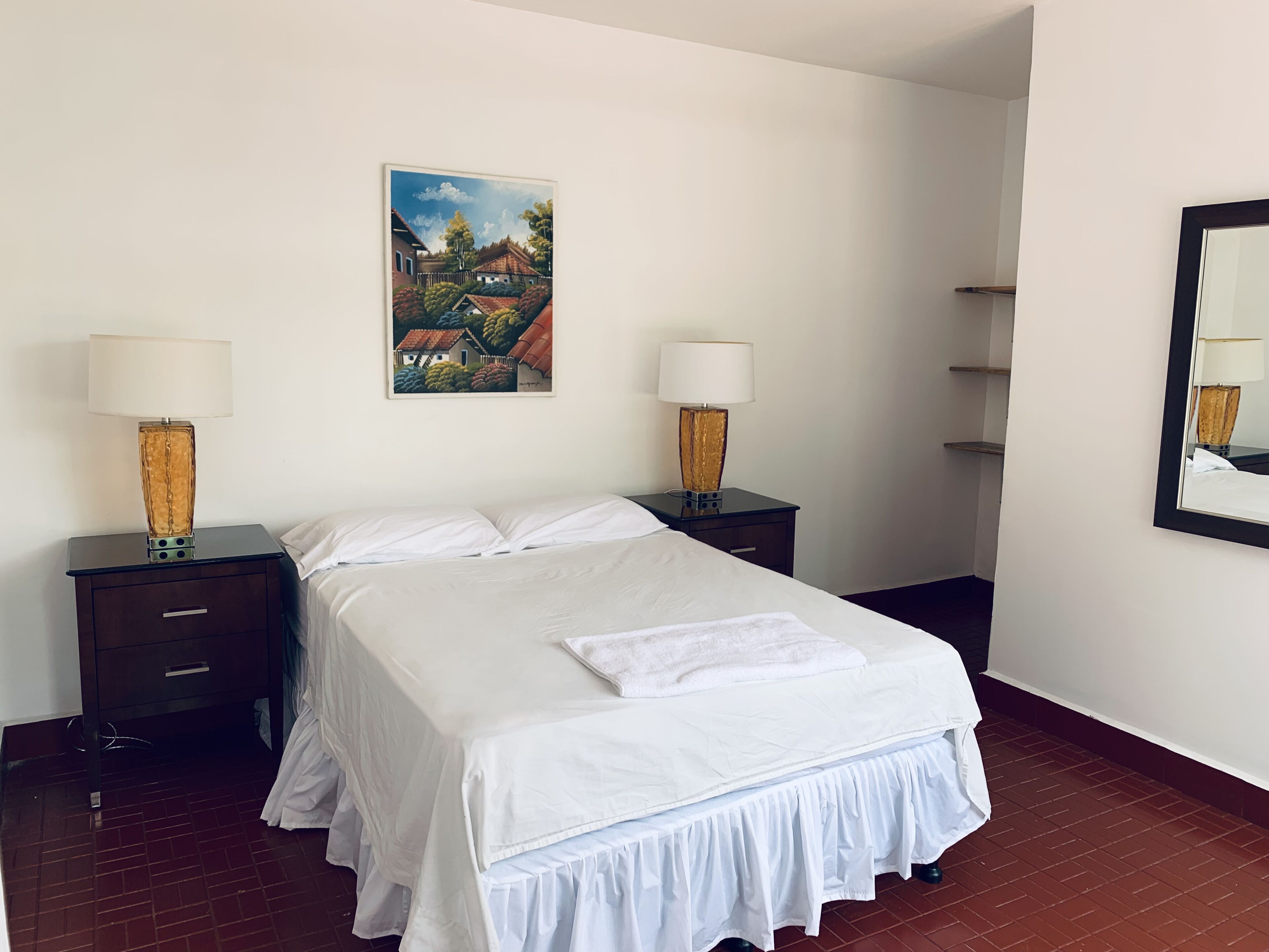 Habitación de hotel con una cama, dos mesitas de noche con lámparas, un cuadro en la pared y un espejo en la pared opuesta.