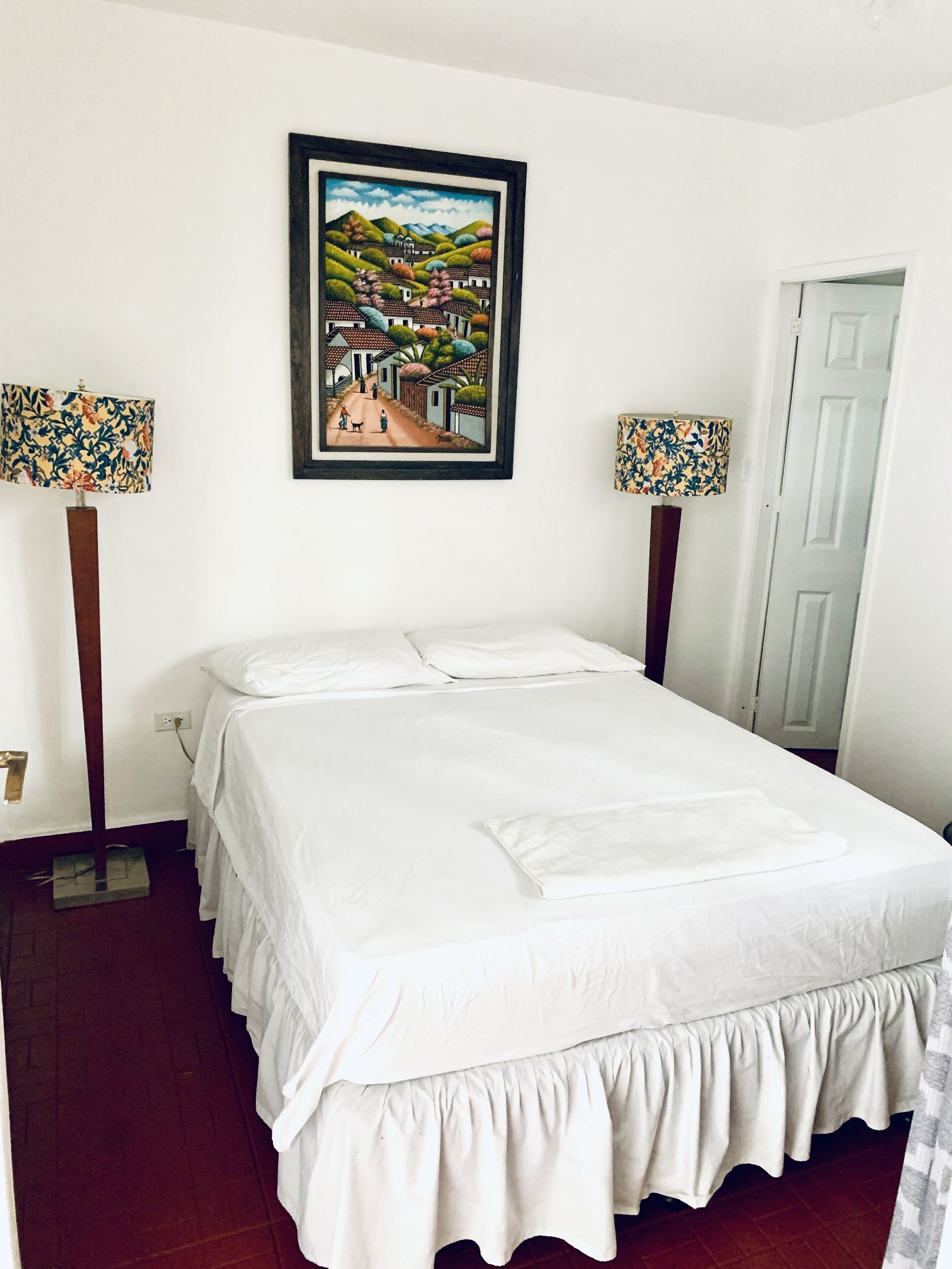 Una cama bien hecha con sábanas blancas, dos lámparas con motivos florales y un cuadro enmarcado con una escena de pueblo en la pared.