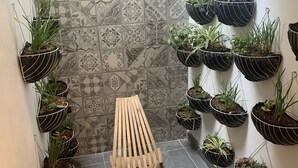 Un couloir étroit avec un mur recouvert de carreaux décoratifs, une chaise en bois et plusieurs pots de fleurs suspendus contenant différentes plantes.