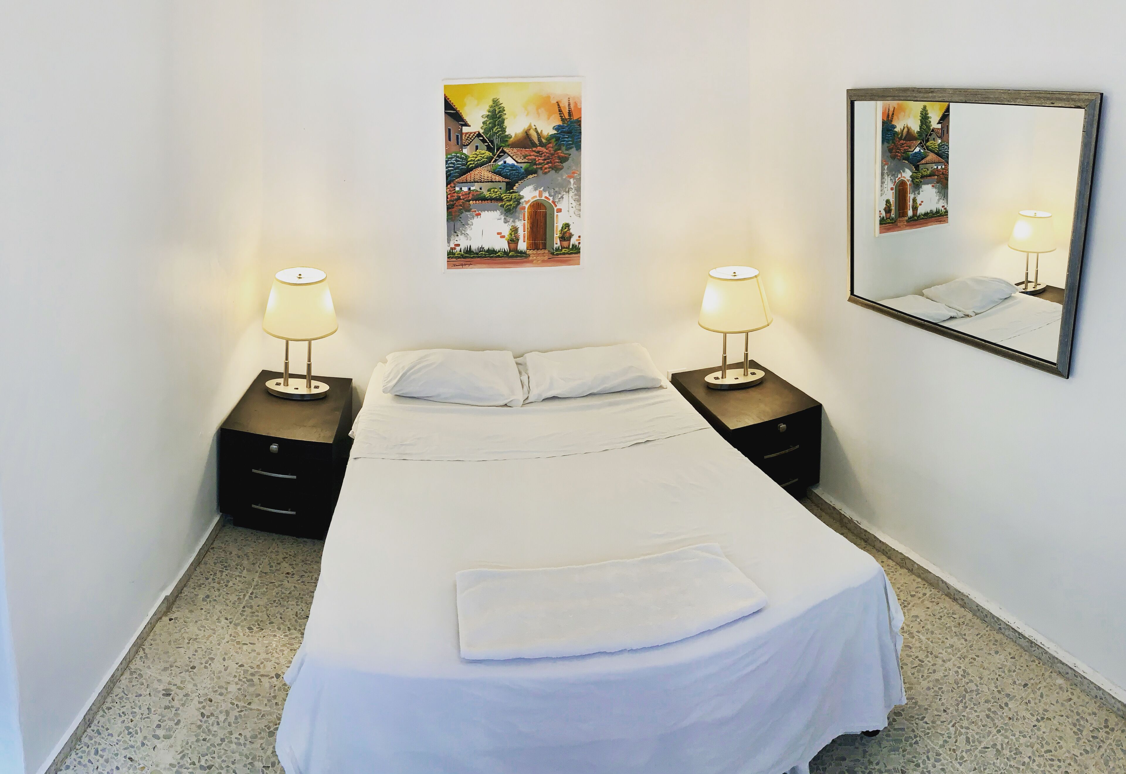 A neatly made bed with white linens, two bedside tables with lamps, and two wall-mounted paintings.