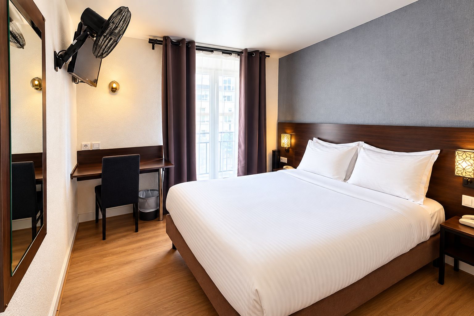 A hotel room with a large bed, a desk, a chair, a mirror, a wall-mounted fan, and a window with curtains.