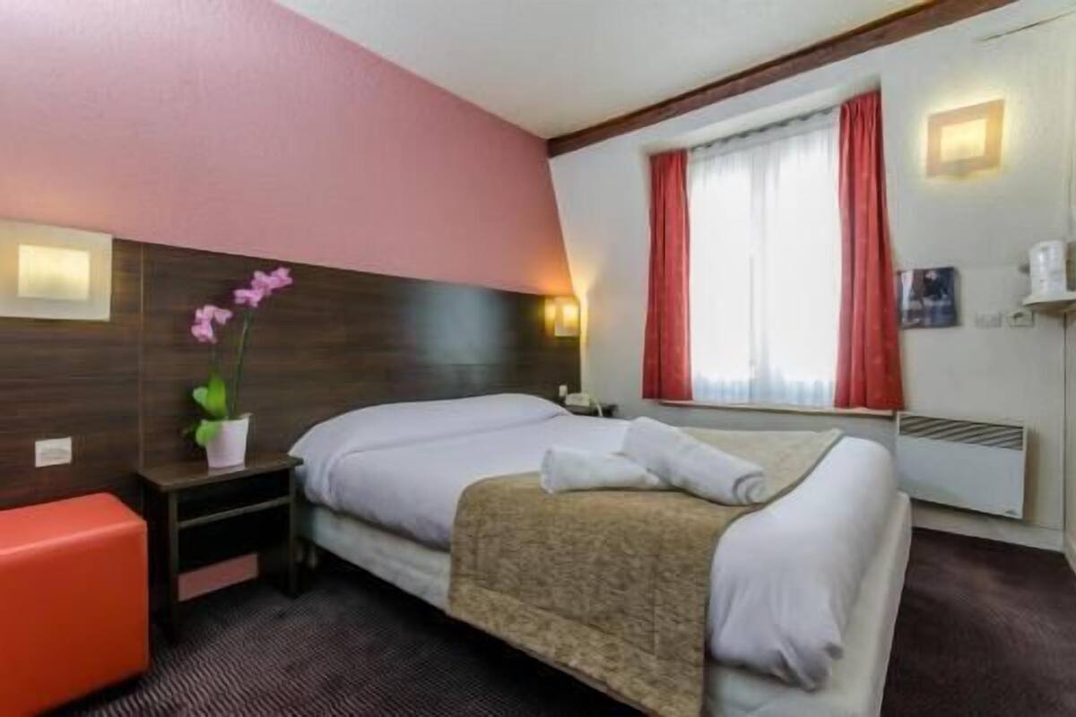 Habitación de hotel con cama, mesita de noche, un banco rojo, planta en maceta y ventana con cortinas.