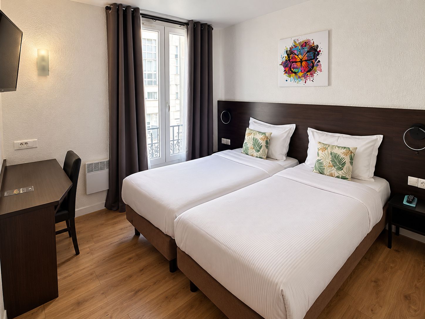 Habitación de hotel con dos camas, un escritorio, una silla, un televisor montado en la pared y una ventana con cortinas.