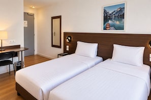 A hotel room with two beds, a desk, a chair, a trash bin, a nightstand, and a wall-mounted mirror. - Hôtel Arc Paris Porte d’Orléans  (Montrouge)