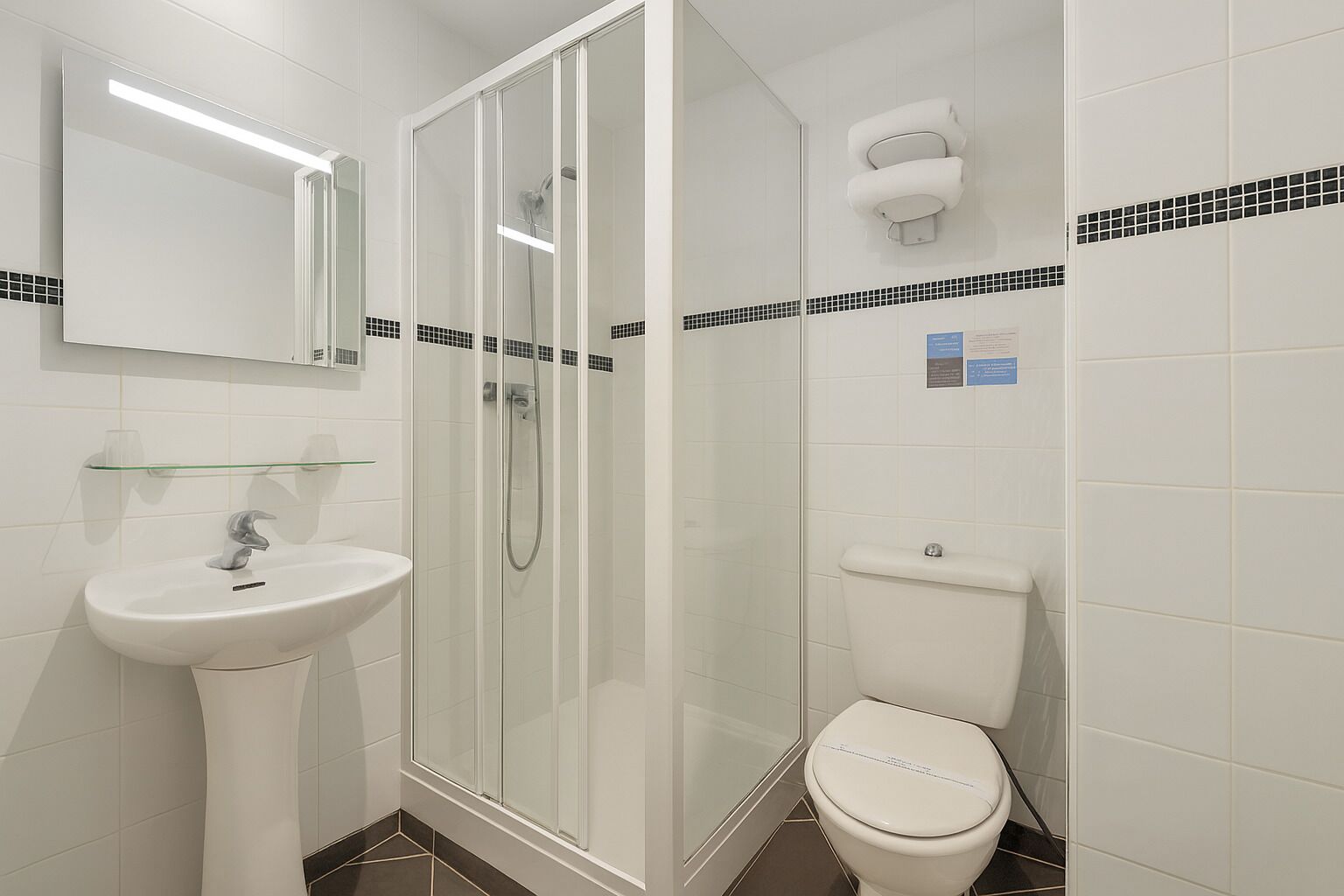 Un baño moderno con ducha envuelta en vidrio, lavamanos pedestal y retrete.