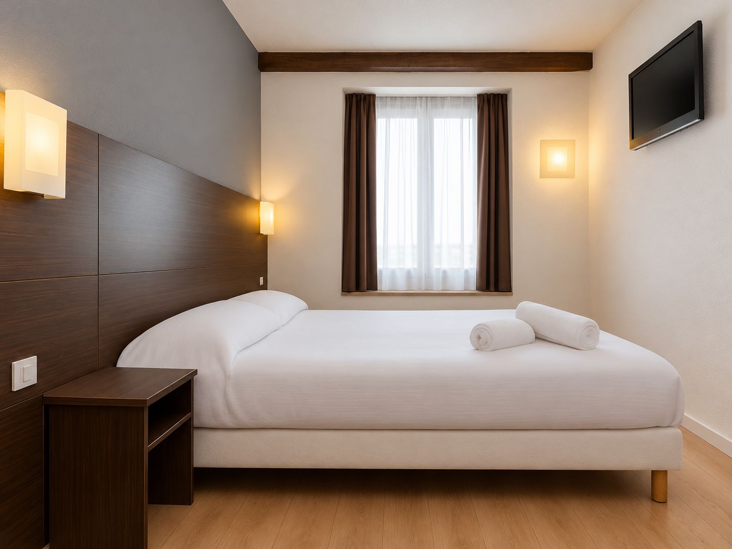 A hotel room with a bed, bedside table, wall-mounted TV, and a window with curtains.