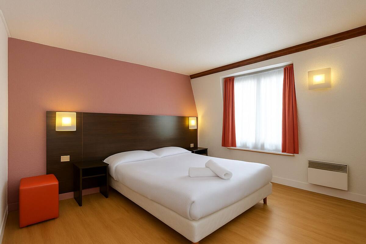 Habitación de hotel ordenada con una cama grande, una mesita de noche, un reposapiés rojo y una ventana con cortinas.