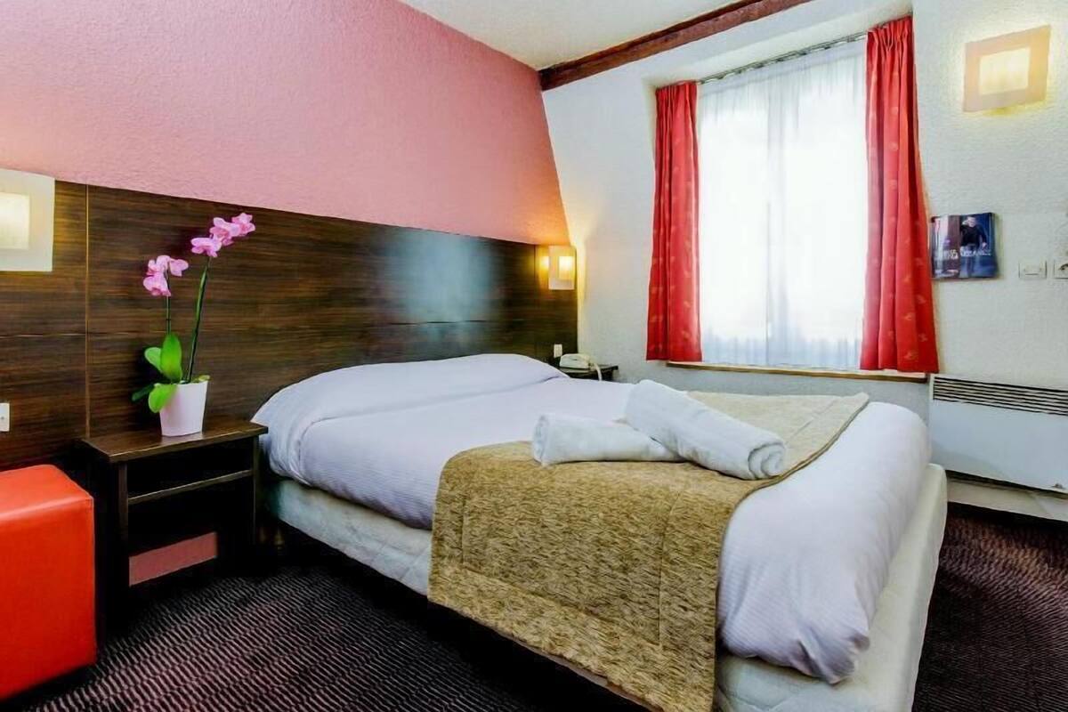 Una habitación de hotel con una cama, una mesita de noche, una silla roja, una orquídea en maceta y una ventana con cortinas rojas.