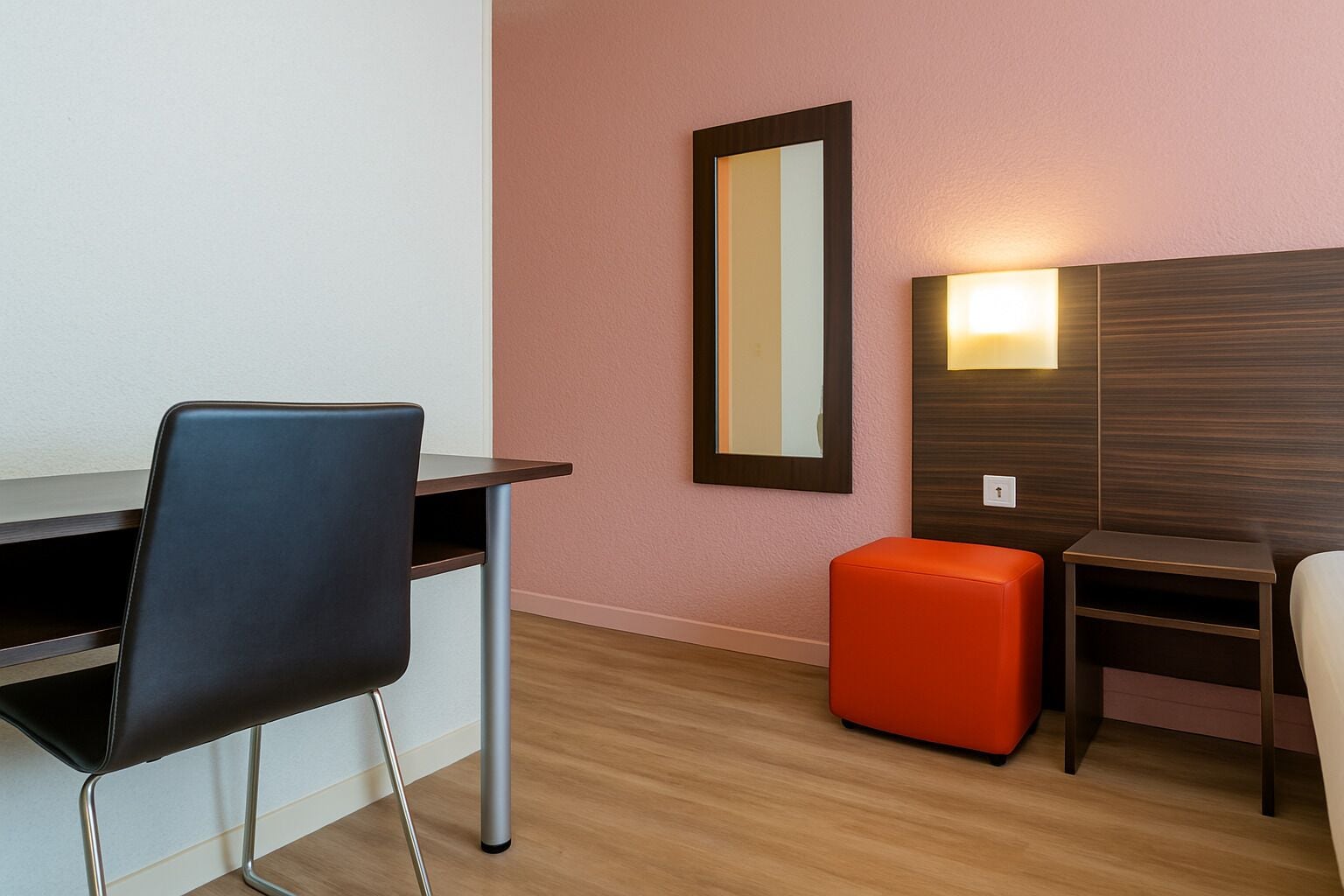 A hotel room with a desk, two chairs, a small orange ottoman, a mirror, and a wall-mounted light fixture.