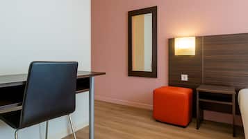 A hotel room with a desk, two chairs, a small orange ottoman, a mirror, and a wall-mounted light fixture.