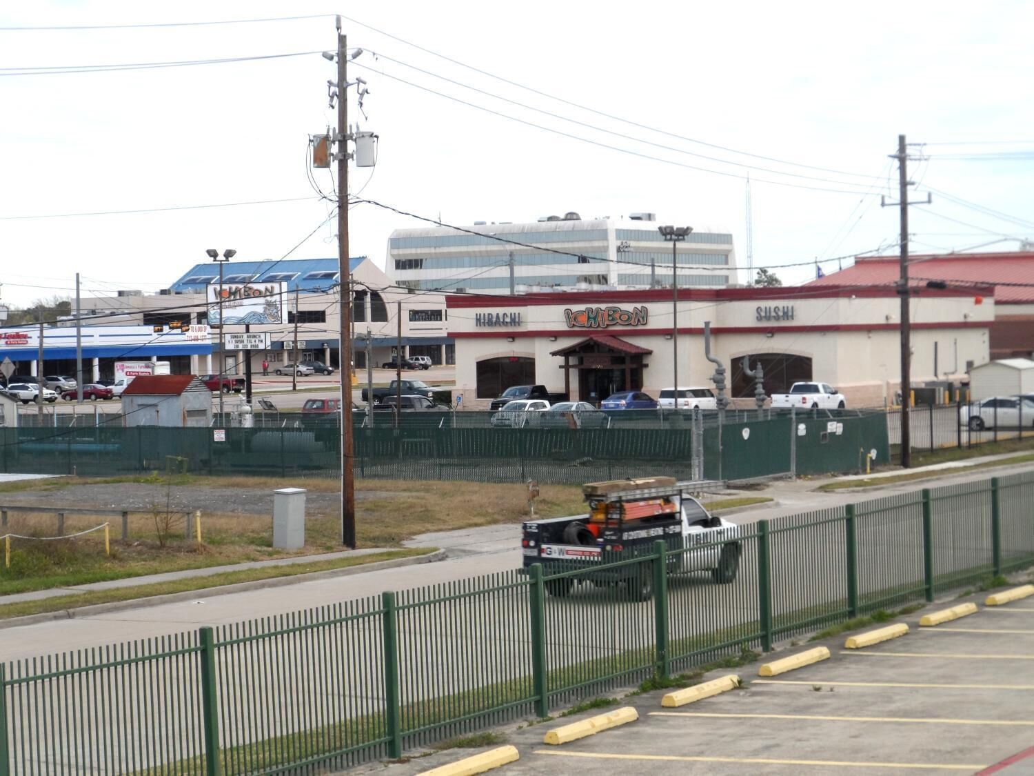 A commercial area with a parking lot, a fenced construction site, and a building with signage for IHBCON and SUSHI.