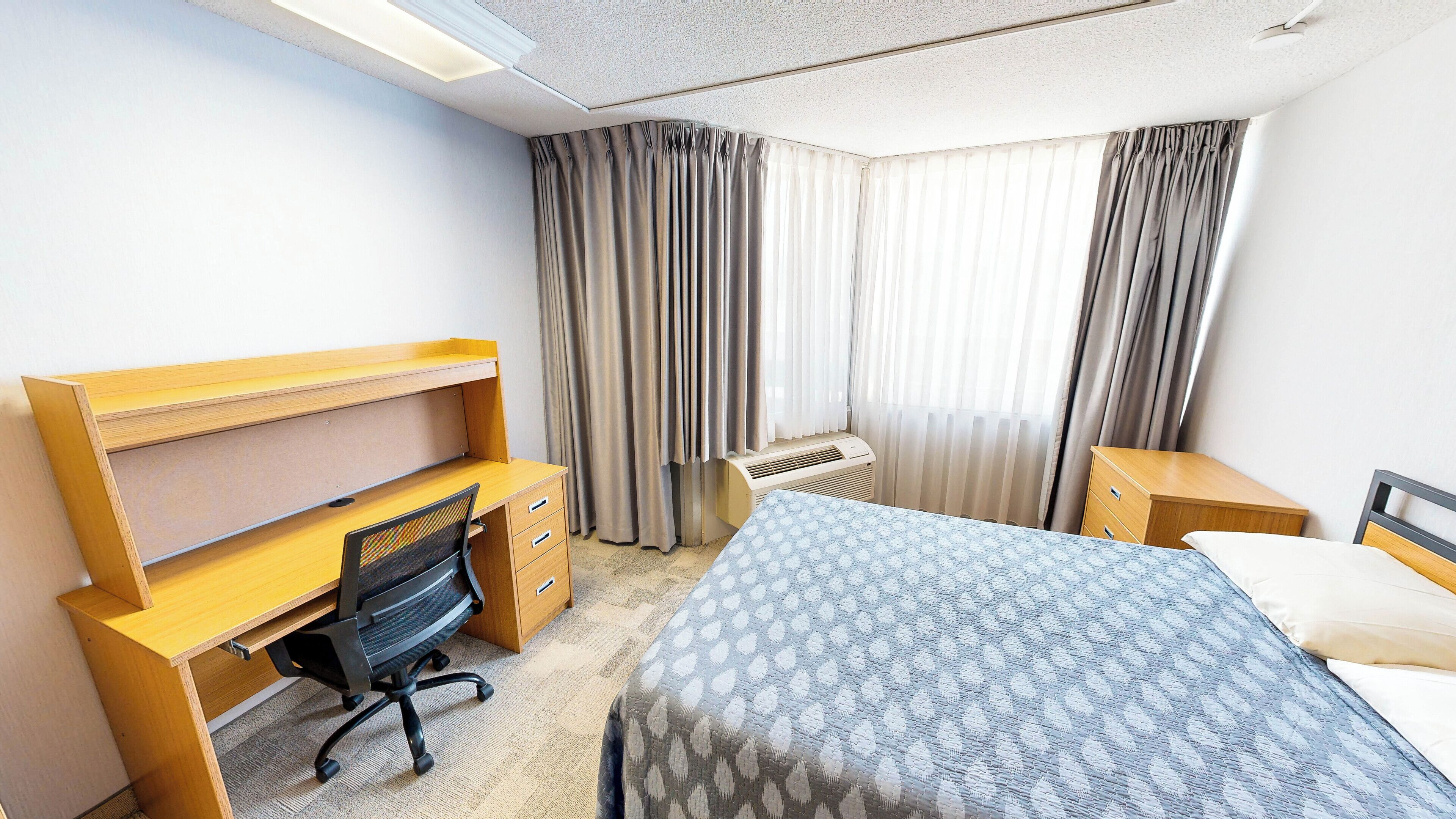 A hotel room with a bed, a desk with a chair, a heater, and curtains.