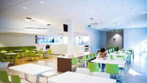 Une cafétéria moderne avec des tables blanches et des chaises vertes, un espace de service central et une vue sur le paysage urbain grâce à de grandes fenêtres.