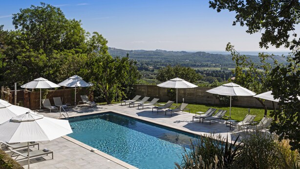 A rectangular swimming pool with lounge chairs and umbrellas, surrounded by trees and a wooden fence, with a view of distant hills. - Le Mas d'Aigret (Les Baux-de-Provence)