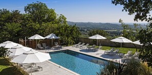 A rectangular swimming pool with lounge chairs and umbrellas, surrounded by trees and a wooden fence, with a view of distant hills. - Le Mas d'Aigret (Les Baux-de-Provence)