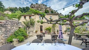 An outdoor dining area with stone walls, tables set for dining, and a view of a stone building with a balcony. - Le Mas d'Aigret (Les Baux-de-Provence)