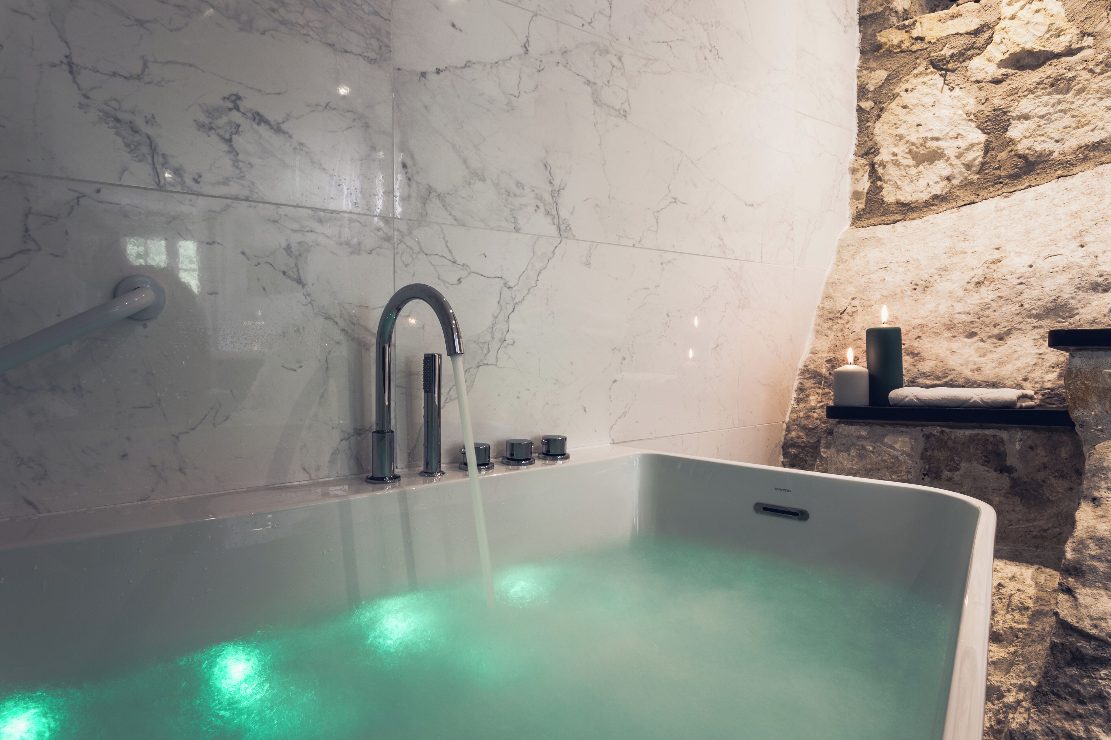 A modern bathroom with a marble wall, a bathtub with green lighting, a stone wall, and a shelf with candles.
