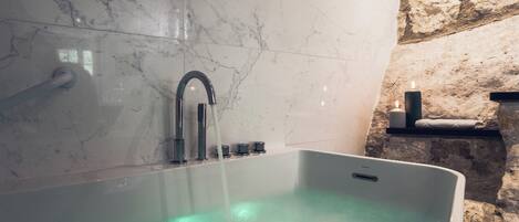 A modern bathroom with a marble wall, a bathtub with green lighting, a stone wall, and a shelf with candles.