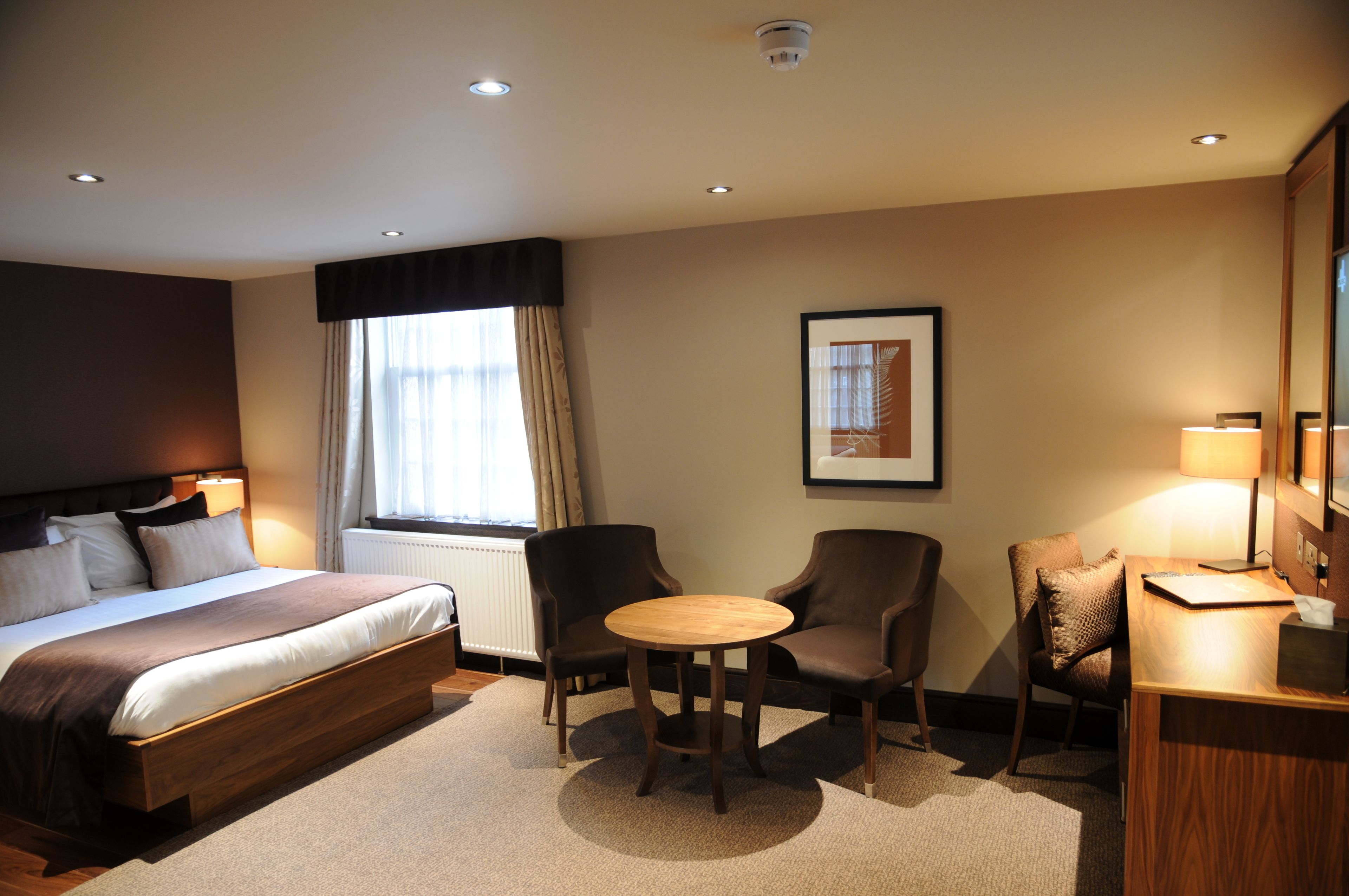 A hotel room with a bed, two armchairs, a small round table, a desk with a lamp, and a framed picture on the wall.