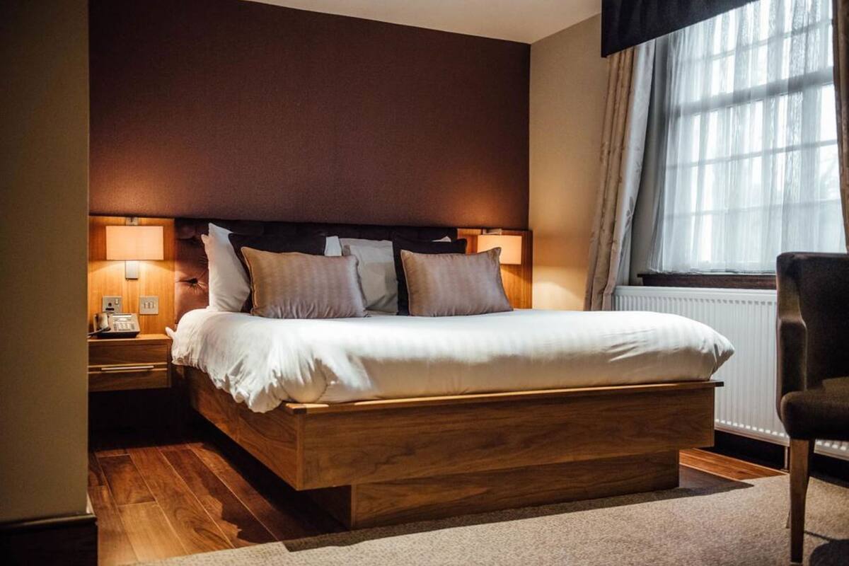 A neatly made bed in a hotel room with a wooden headboard, a nightstand with a lamp, and a window with curtains.