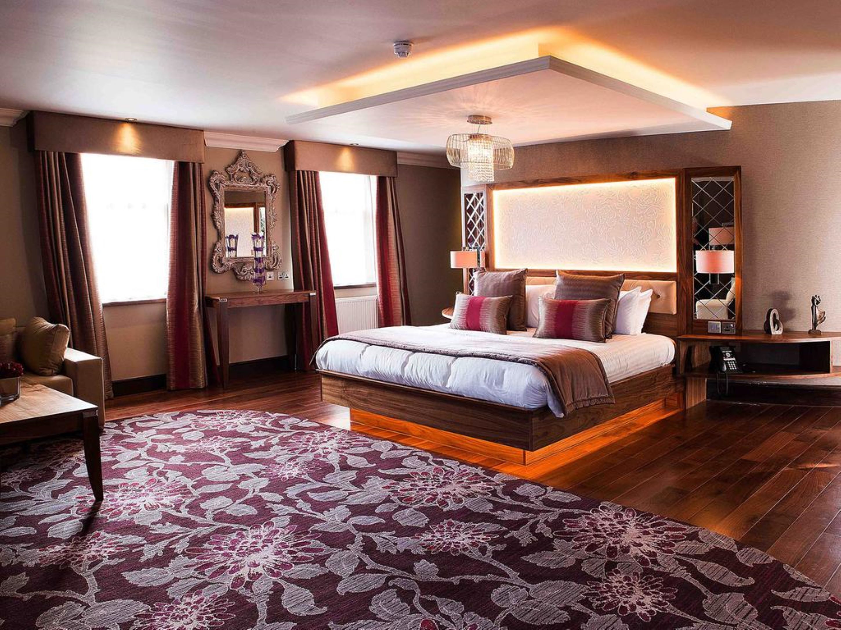 A spacious bedroom with a large bed, a wooden nightstand, a desk, and a chair. There is a large window with curtains, a mirror, and a chandelier.