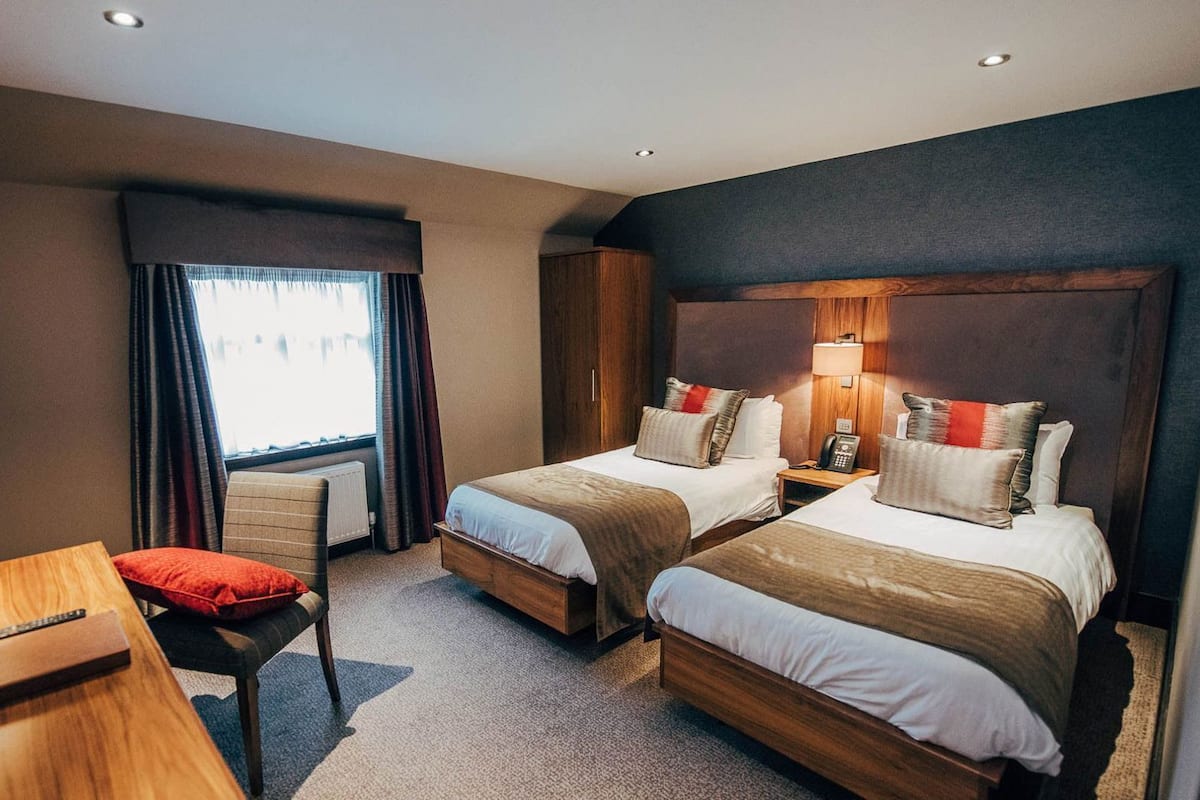 A hotel room with two beds, a desk, a chair, a lamp, and a window with curtains.