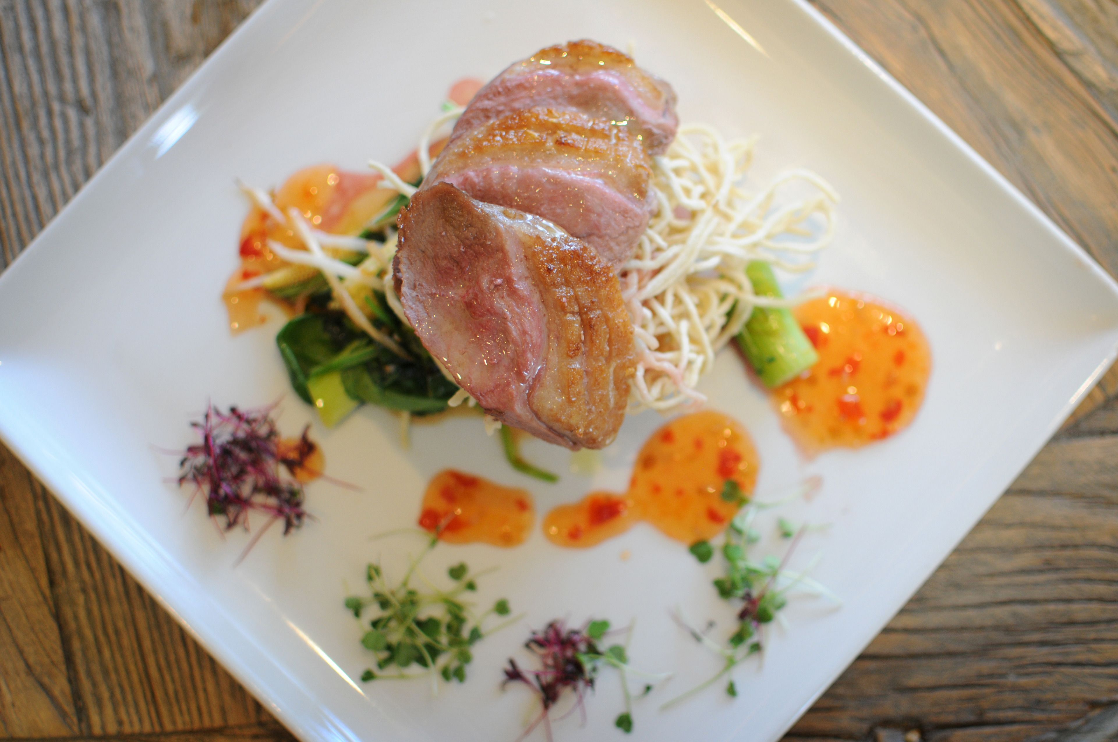 A square plate with a piece of meat, vegetables, and sauce.