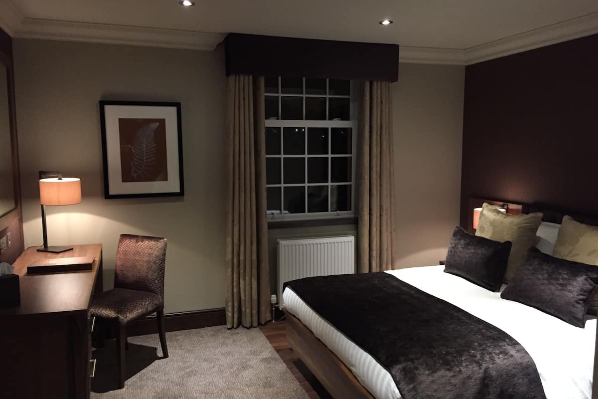 A hotel room with a bed, a desk, a chair, a window with curtains, and a framed picture on the wall.