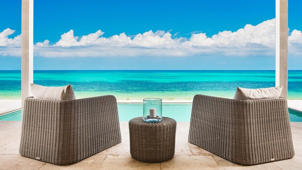 A wicker sofa with cushions and a glass candle holder in front of a pool, overlooking a turquoise sea and clear sky.