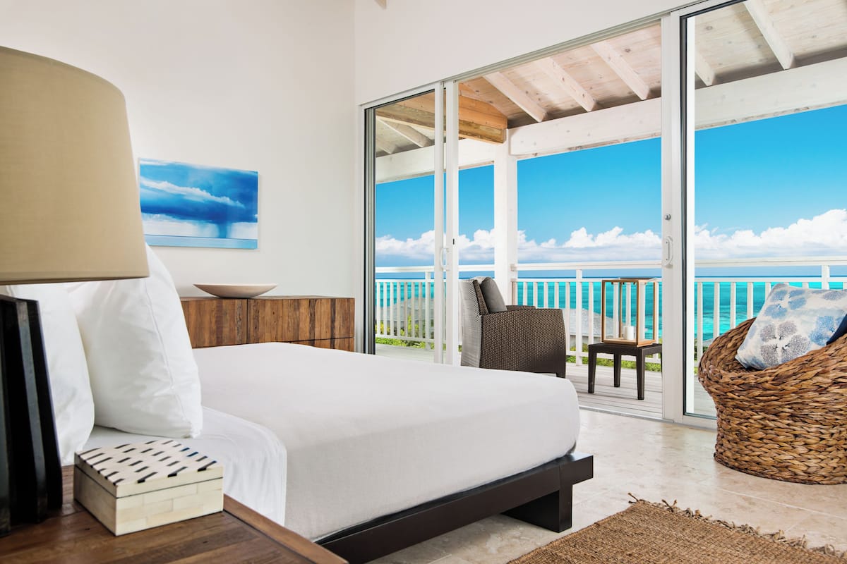 A bedroom with a large bed, a wooden nightstand, a bedside table, a lamp, a wall-mounted painting, and a view of the ocean through a sliding glass door.