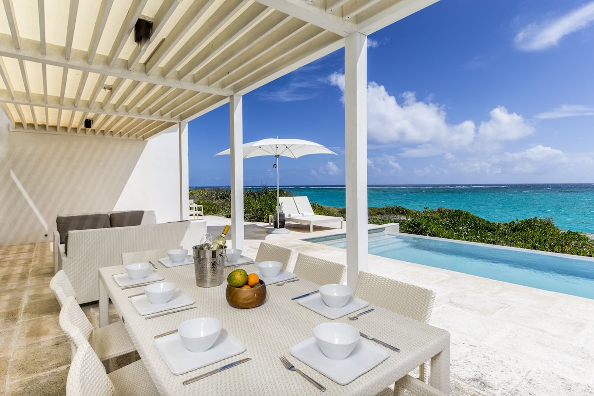 A covered outdoor dining area with a view of the ocean, white furniture, and a table set for a meal.