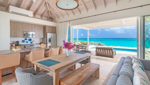 A modern kitchen with wooden cabinets, a dining area with a wooden table and benches, and a large sliding glass door leading to a balcony with a view of the ocean.
