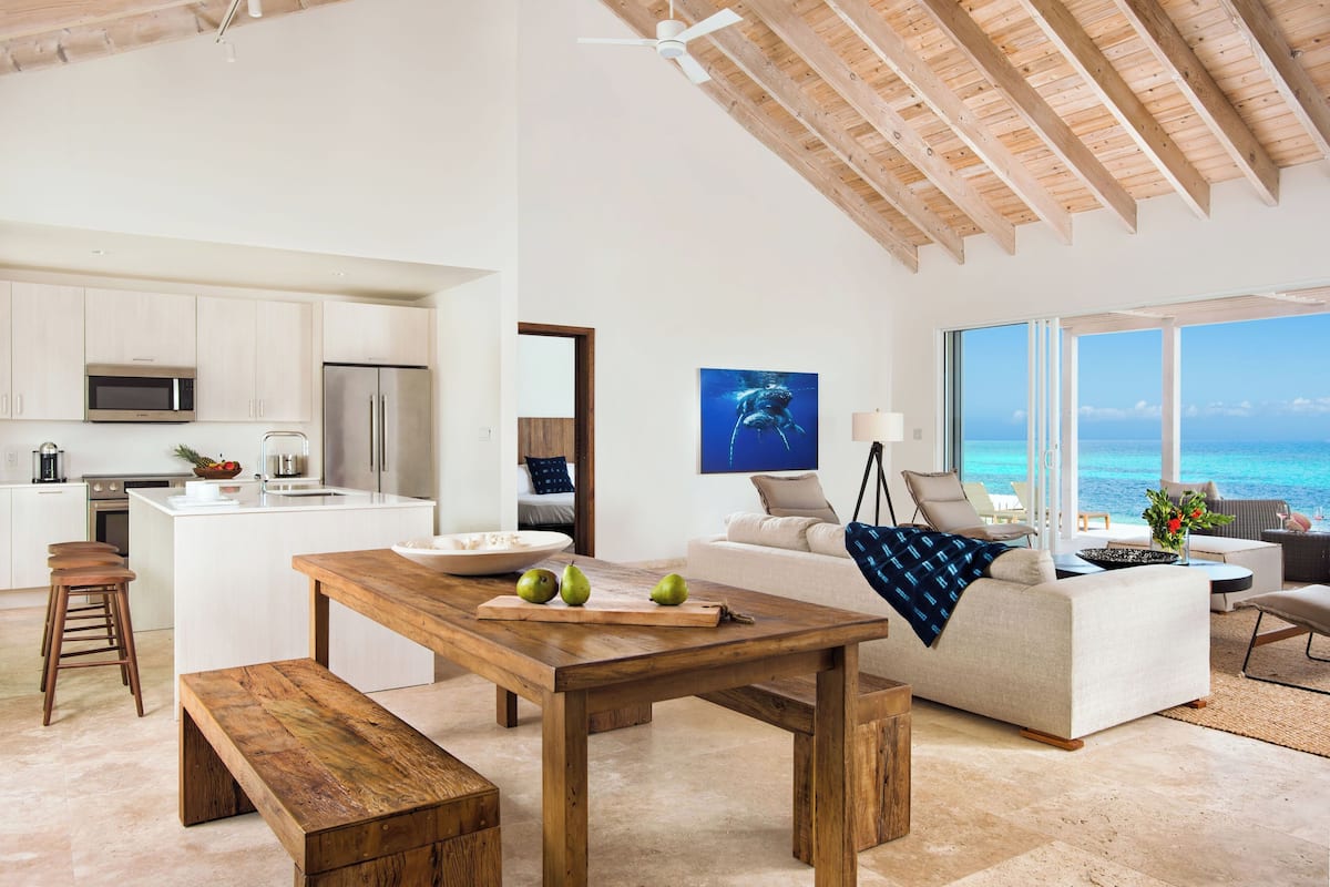 A modern kitchen with white cabinets, a wooden dining table, and a bench. There is a large window with a view of the ocean.