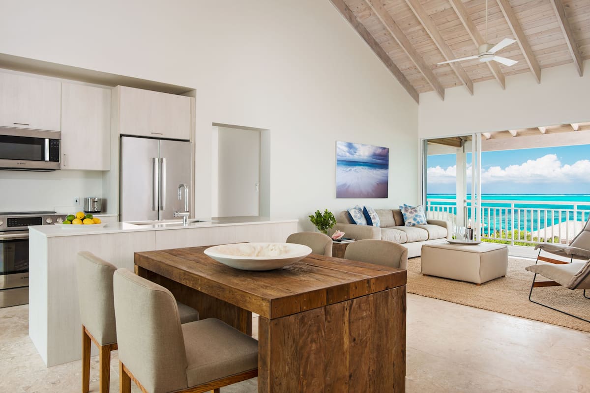 A modern kitchen with white cabinets, a wooden dining table, and a large island. There is a view of the ocean through a balcony.