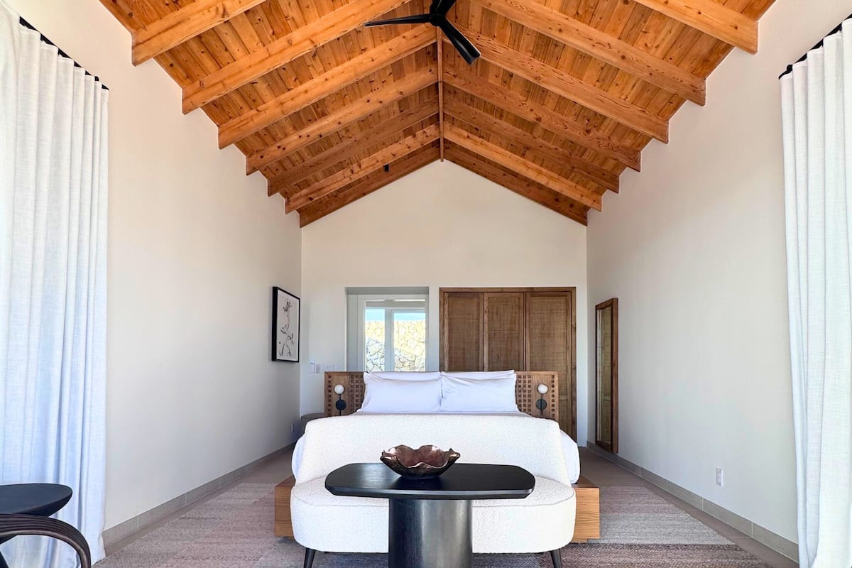 A spacious bedroom with a high wooden ceiling, a large bed, a bedside table, and a view of the outside.
