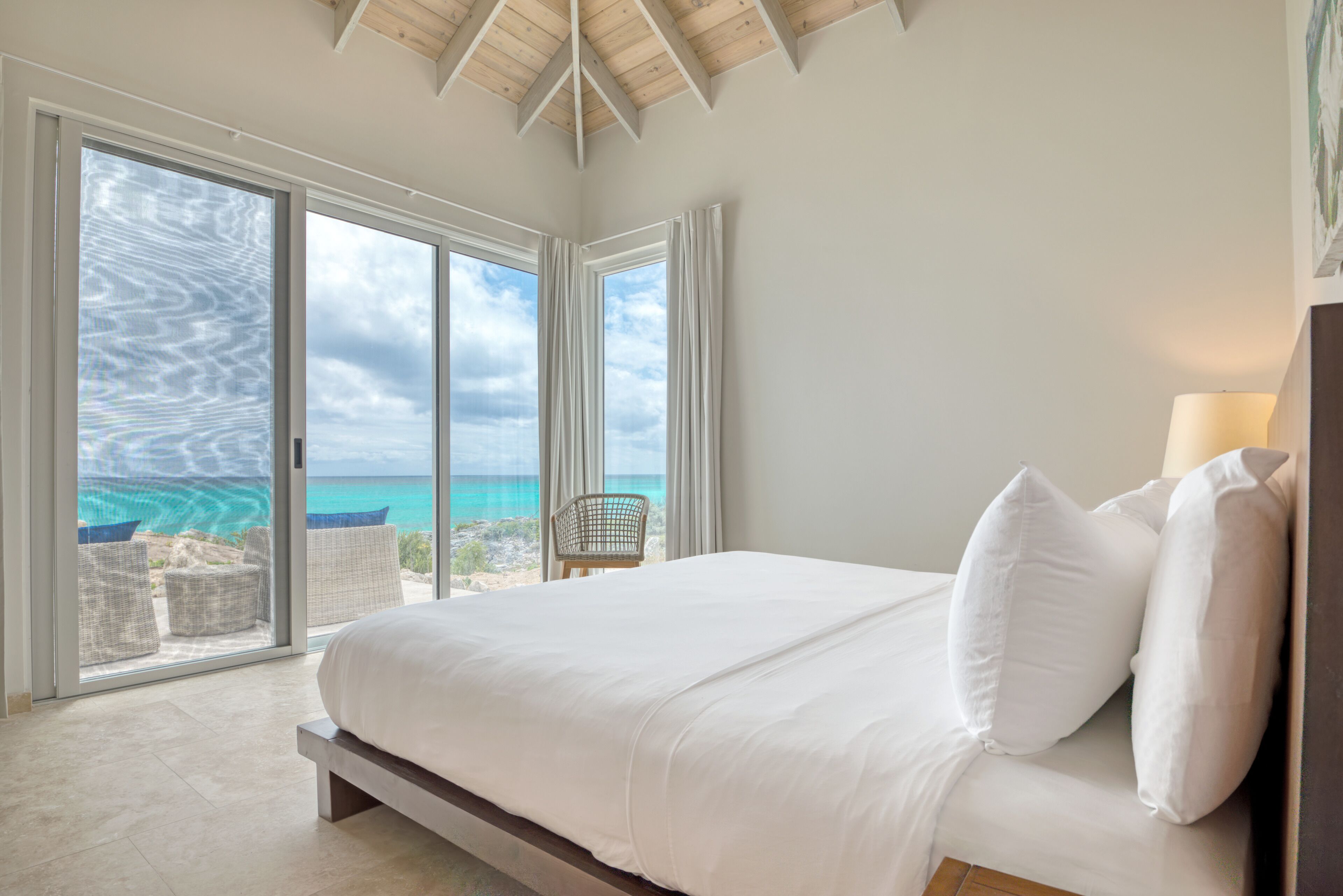 A bedroom with a large bed, white bedding, and a view of the ocean through floor-to-ceiling windows.