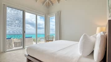 A bedroom with a large bed, white bedding, and a view of the ocean through floor-to-ceiling windows.