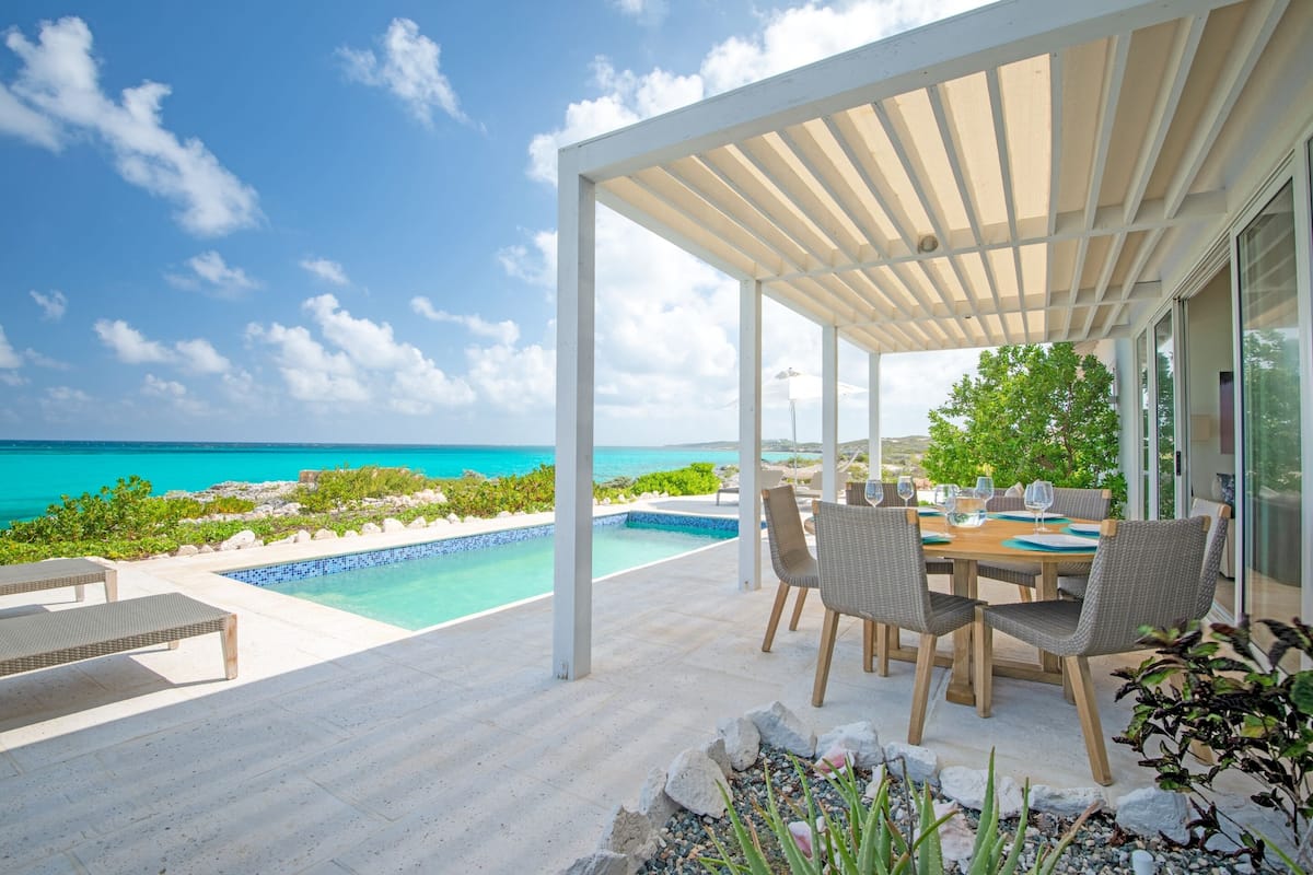 A covered outdoor area with a pool, lounge chairs, and a table set for dining, overlooking a turquoise sea.