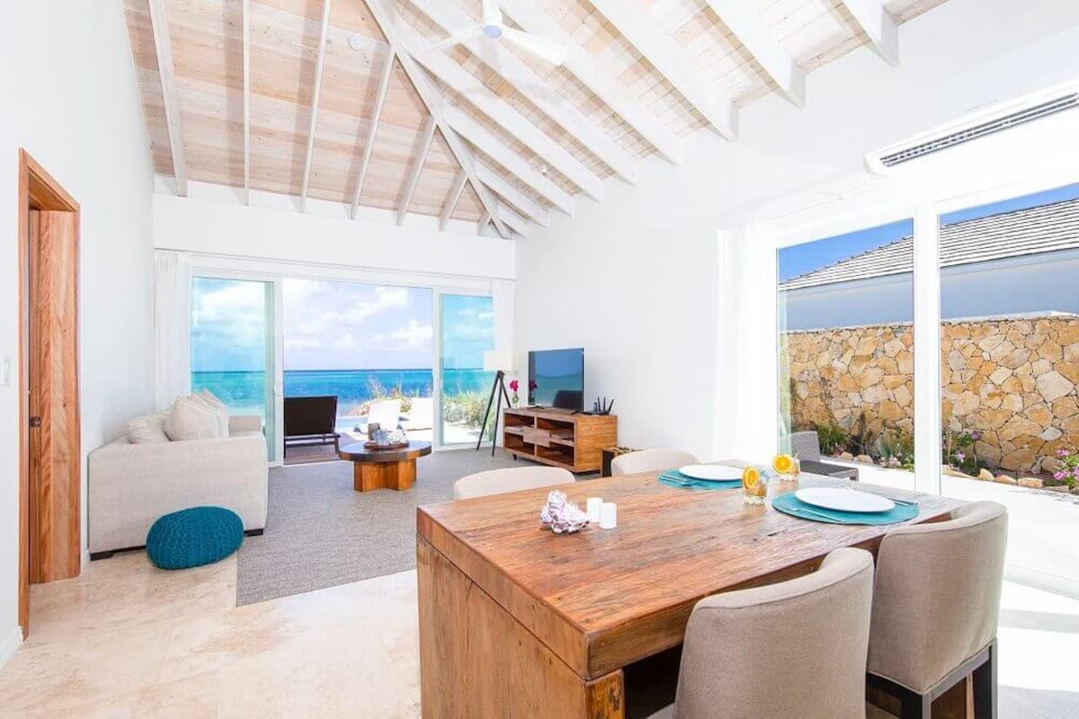 A spacious living room with a wooden dining table, comfortable seating, a television, and a view of the ocean.