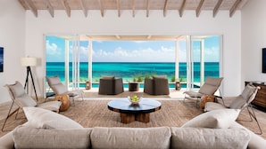 A spacious living room with a large window overlooking a turquoise ocean. There are two sofas, a coffee table, and two armchairs.