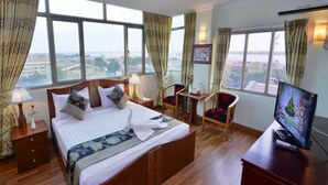 Habitación de hotel con una cama grande, un escritorio con televisor, dos sillas, una mesita y vistas a la ciudad a través de amplias ventanas.