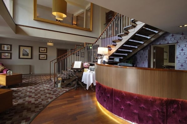 Vestíbulo de hotel con mostrador de recepción de madera, una escalera y una alfombra estampada.