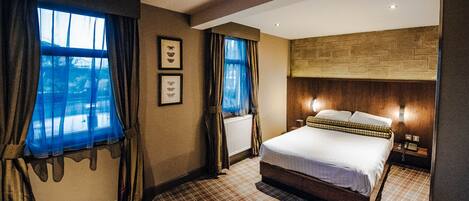 A hotel room with a large bed, two bedside tables, a wooden headboard, and a view of the outdoors through the windows.