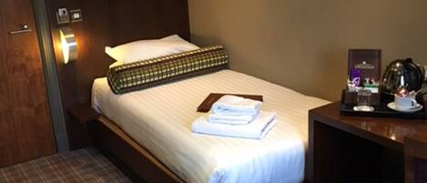 A hotel room with a bed, a bedside table with a lamp, a trash bin, and a wall-mounted light fixture.