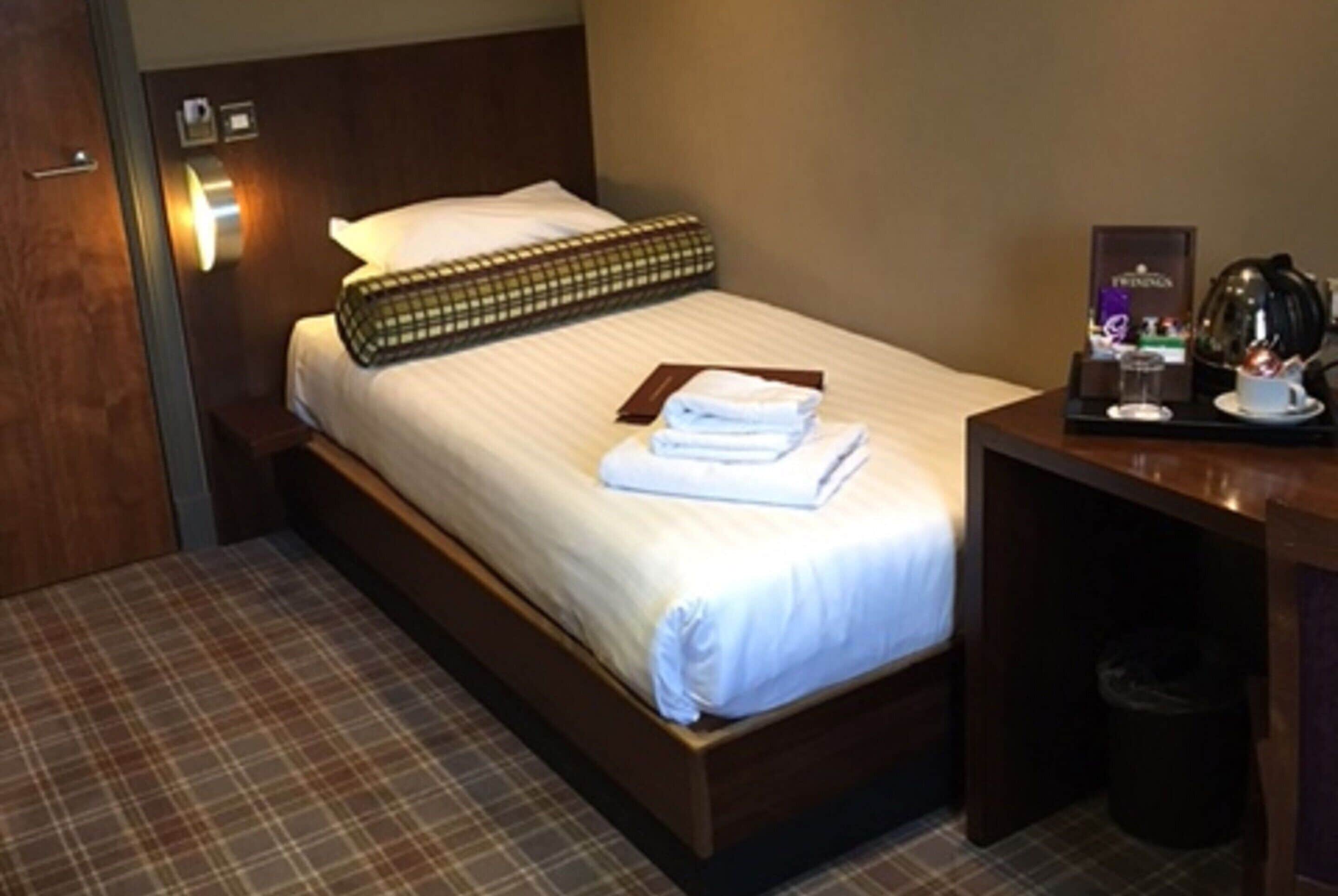 Een hotelkamer met een bed, een nachtkastje met een lamp, een kleine prullenbak en een koffiezetapparaat op de tafel.