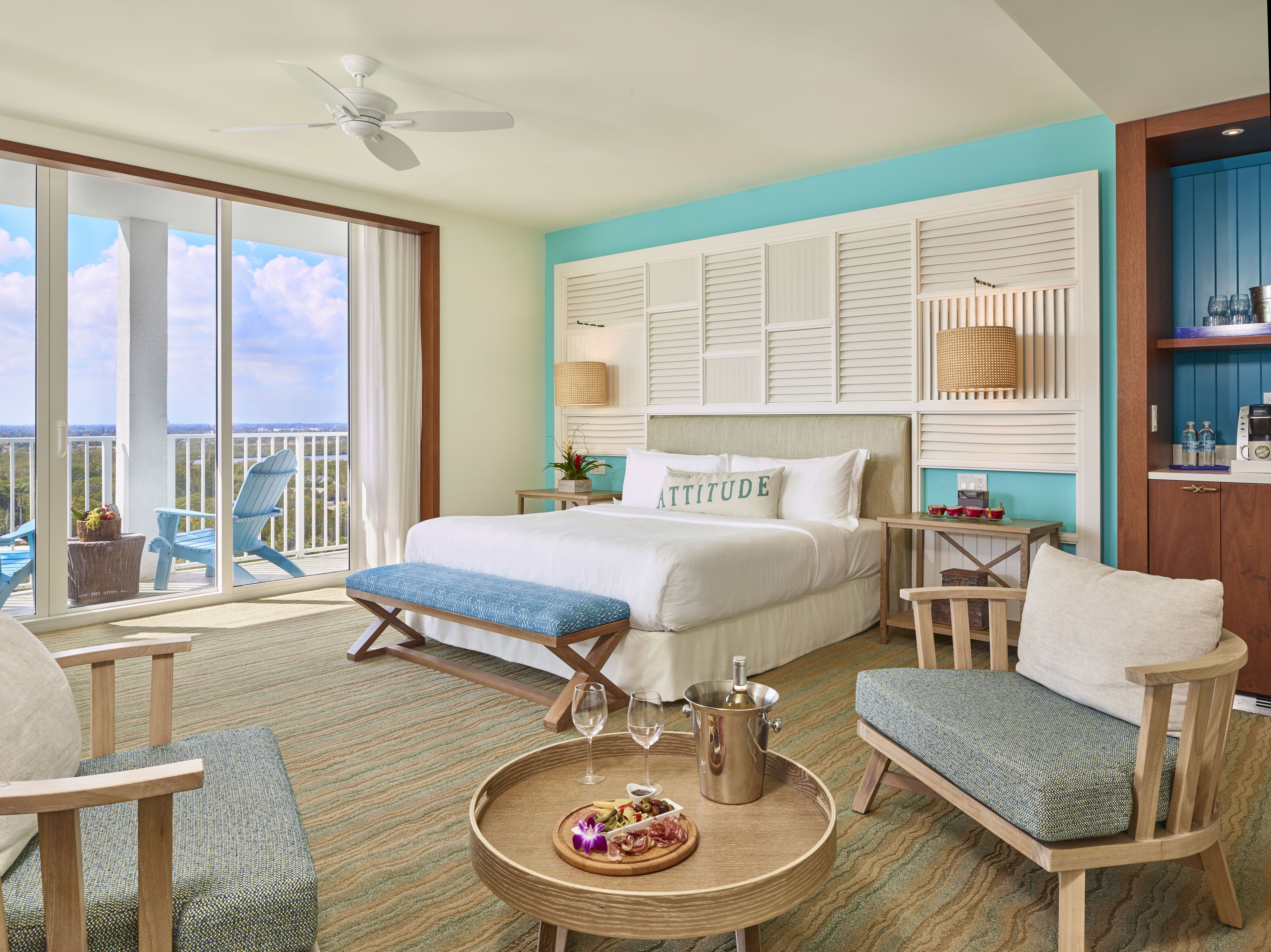 Une chambre d’hôtel avec un lit, un balcon avec des chaises, une table à manger avec une bouteille et des verres, et un minibar.