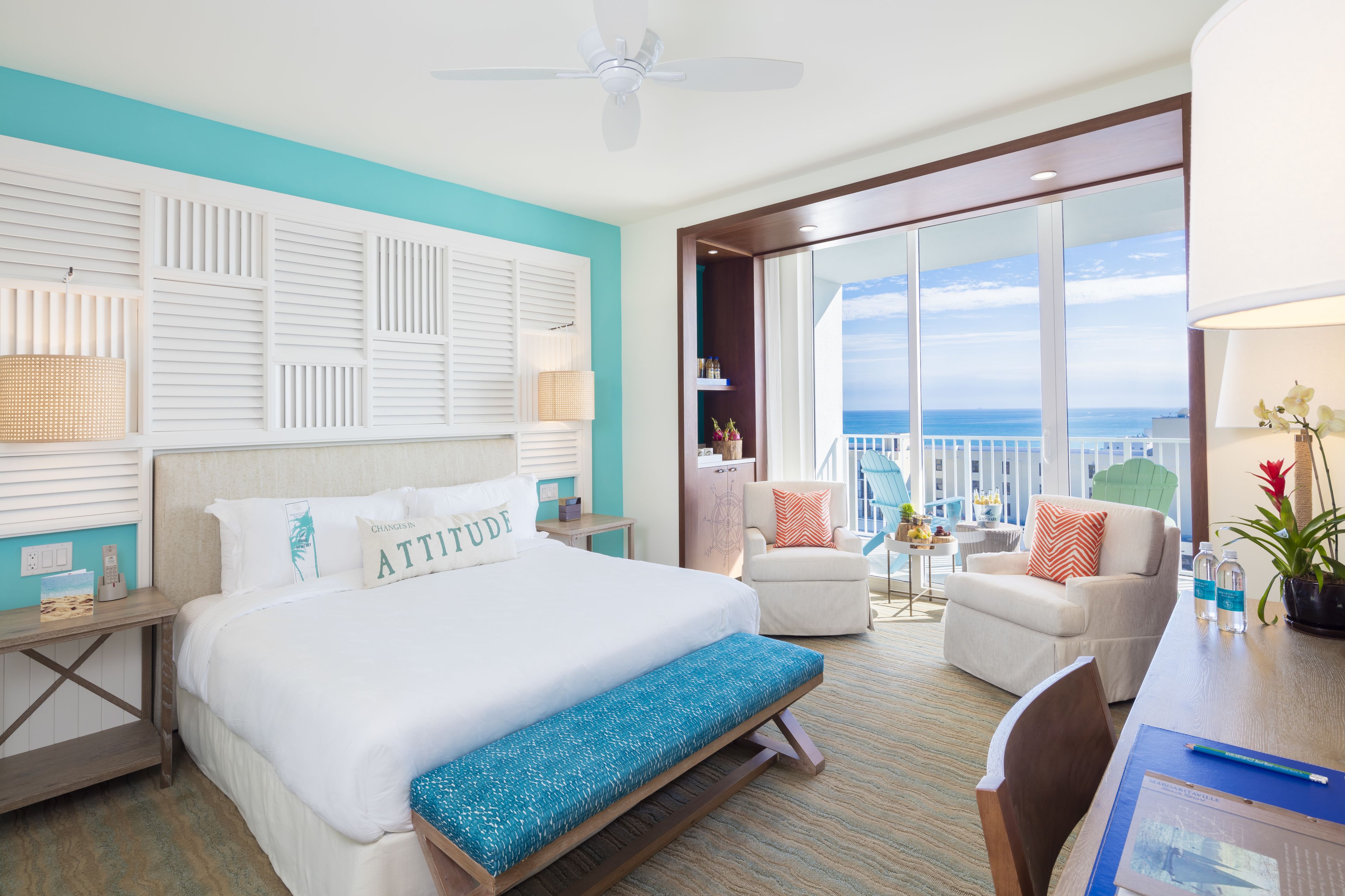 Ein Hotelzimmer mit einem großen Bett, einem Schreibtisch mit Stuhl, einem Balkon mit Meerblick und einem Deckenventilator.