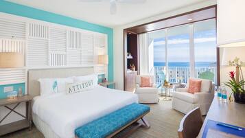 A hotel room with a large bed, a desk with a chair, a balcony with a view of the ocean, and a ceiling fan.